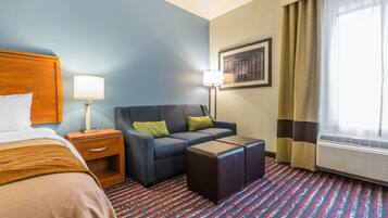 Habitación de hotel con cama, sofá, mesita, mesita de noche, lámpara, aire acondicionado y ventana con cortinas.