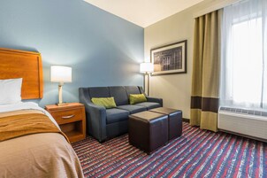 Habitación de hotel con cama, sofá, mesita, mesita de noche, lámpara, aire acondicionado y ventana con cortinas.