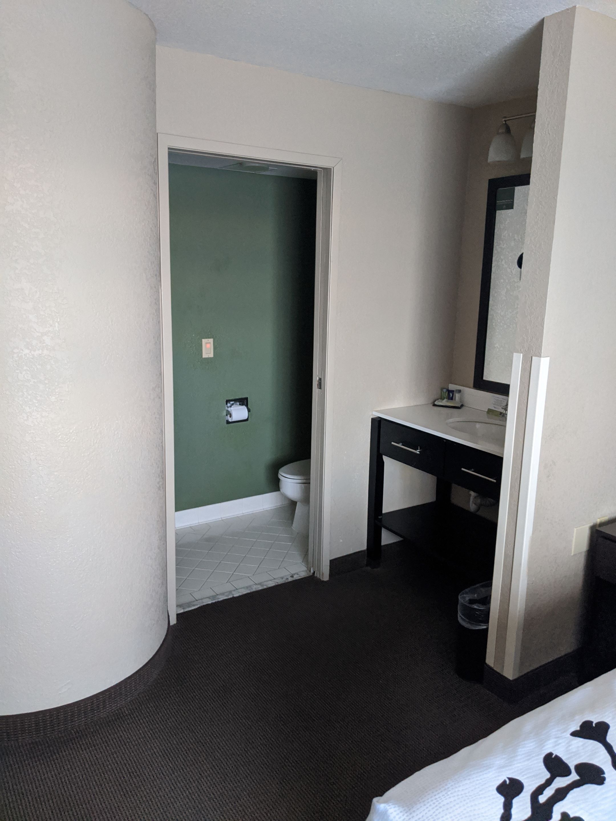 A hotel room with a green wall, a white door leading to a bathroom, a black vanity with a mirror, and a white bed with a patterned blanket.