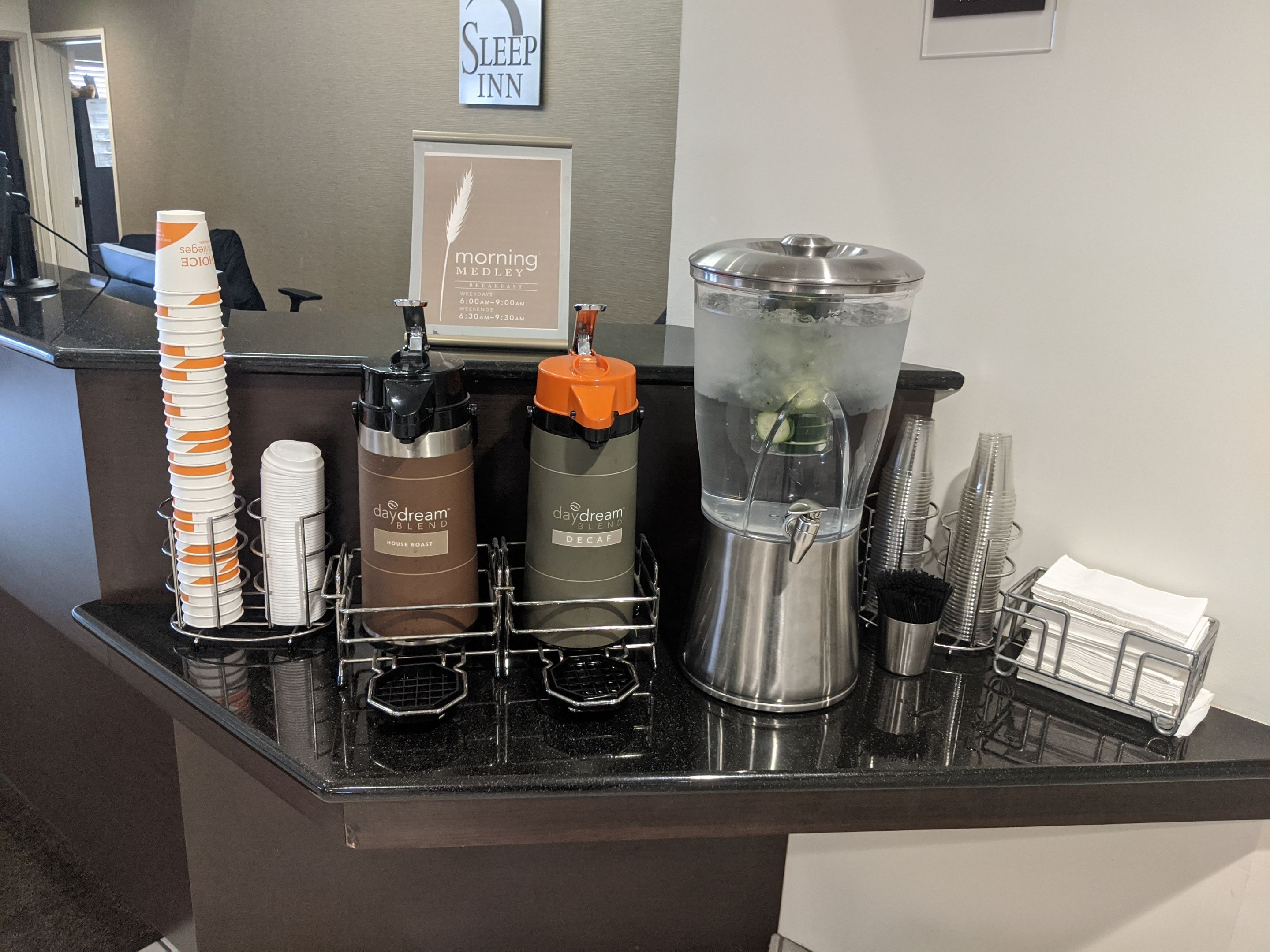 A hotel reception area with a coffee station, a sign for 'Morning Medley Breakfast', and a 'Sleep Inn' sign on the wall.