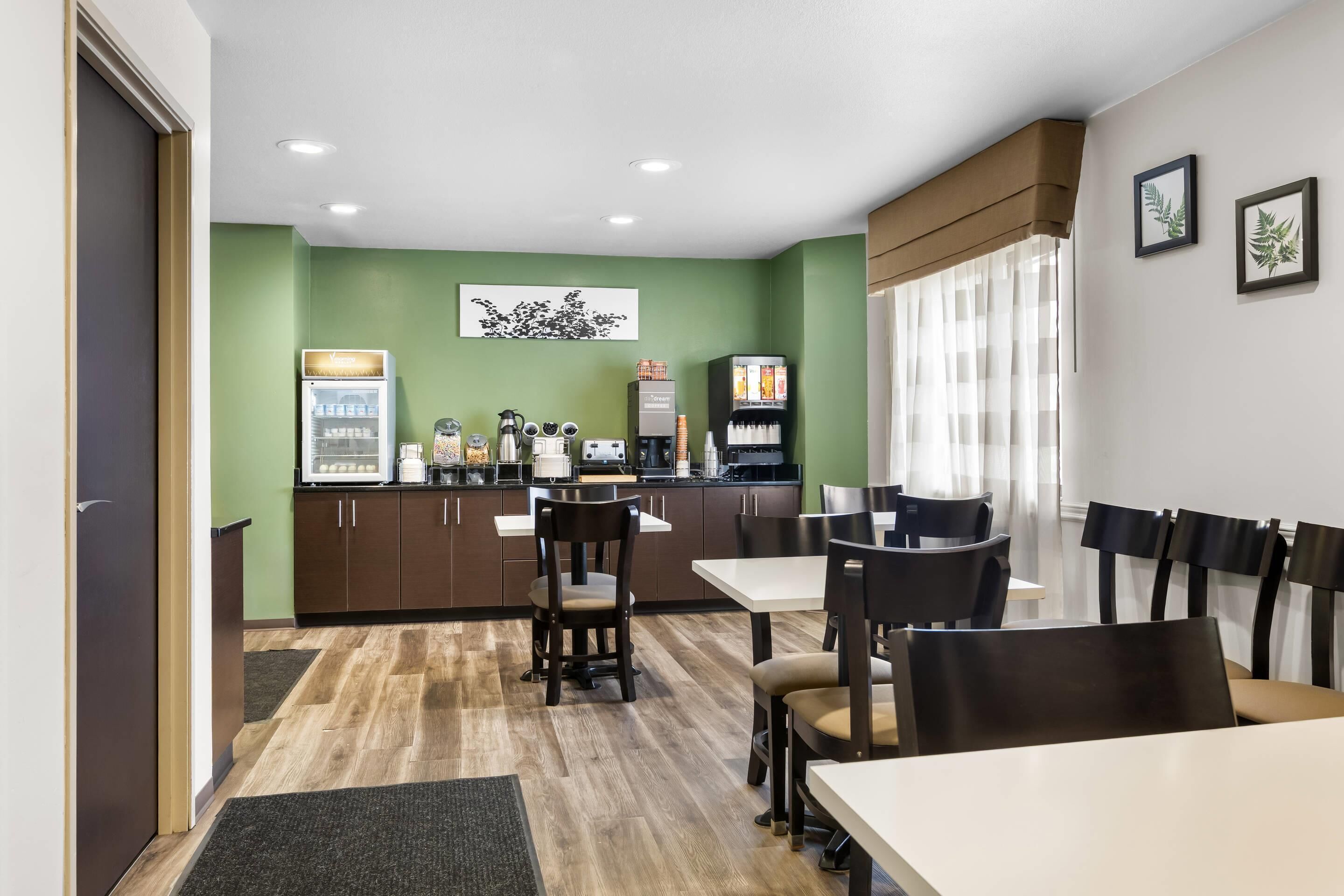 A modern dining area with a kitchen in the background, featuring a refrigerator, coffee machine, and a menu board.
