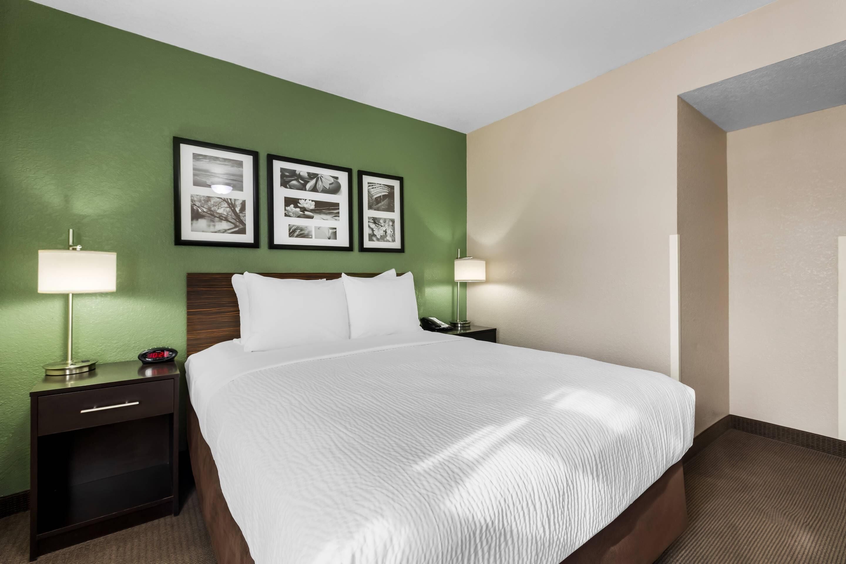 Una habitación de hotel con una cama grande, dos mesitas de noche, una lámpara de noche, un reloj en la pared y tres cuadros enmarcados.
