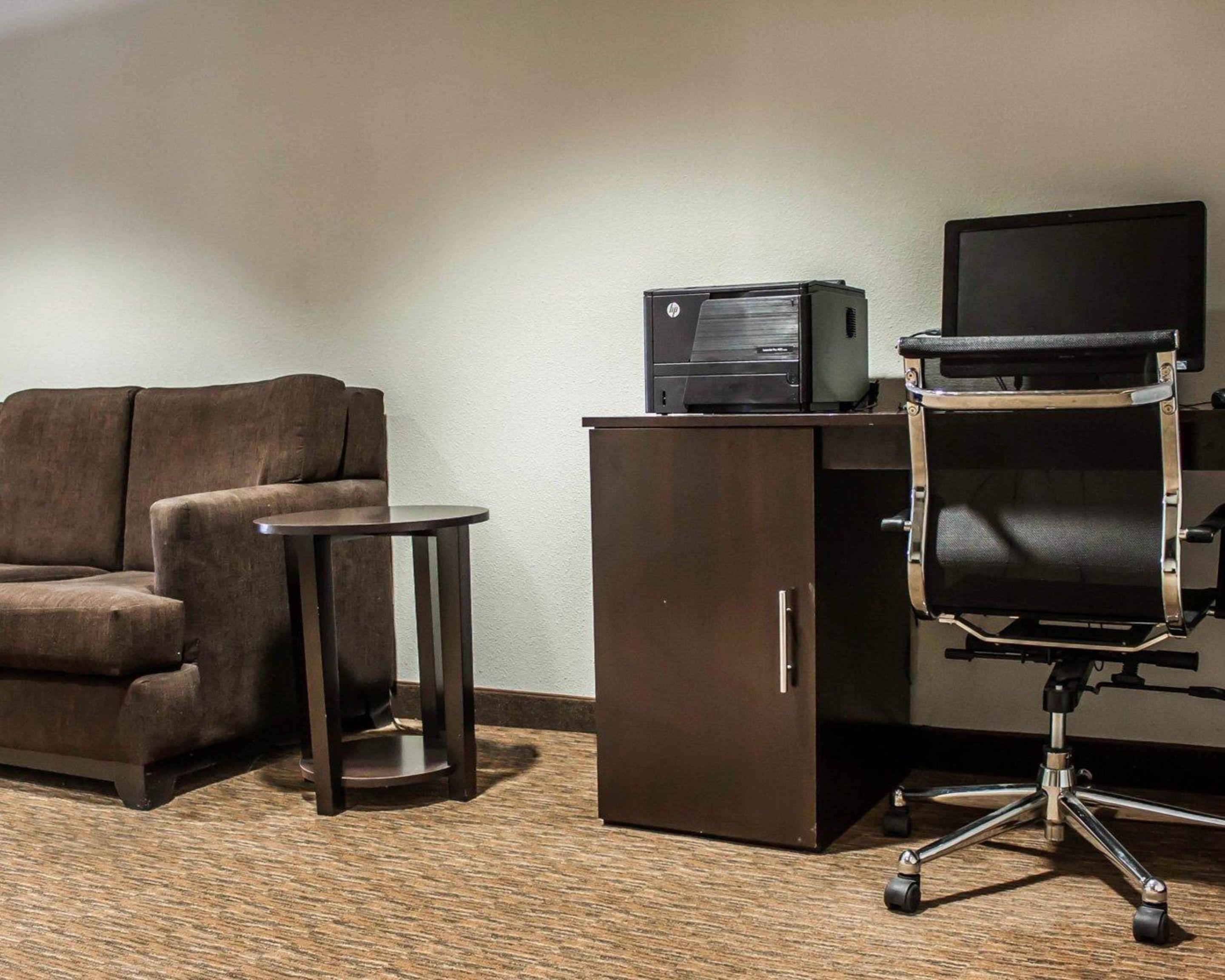 A hotel room with a brown sofa, a small round table, a desk with a computer and printer, and a black office chair.
