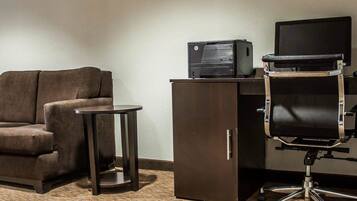 A hotel room with a brown sofa, a small round table, a desk with a computer and printer, and a black office chair.