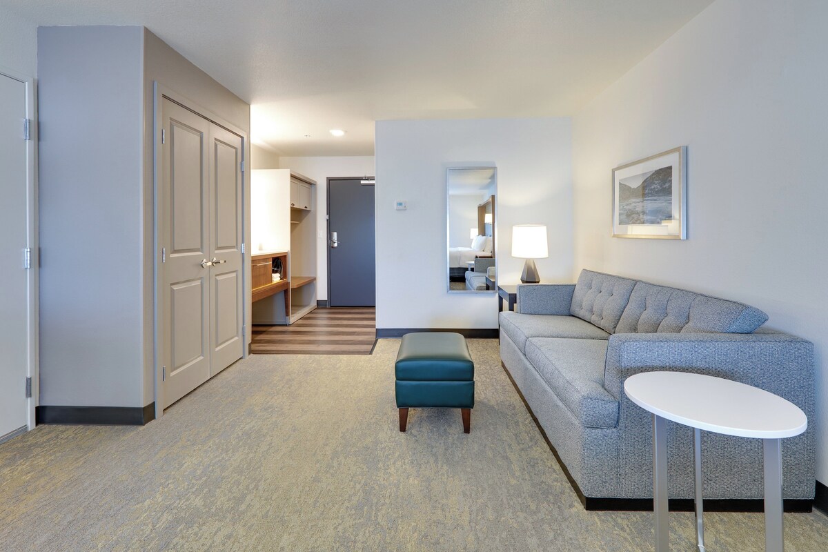 Une chambre d’hôtel avec un canapé gris, une petite table blanche, un pouf turquoise, un miroir et une porte donnant sur un couloir au plancher de bois.