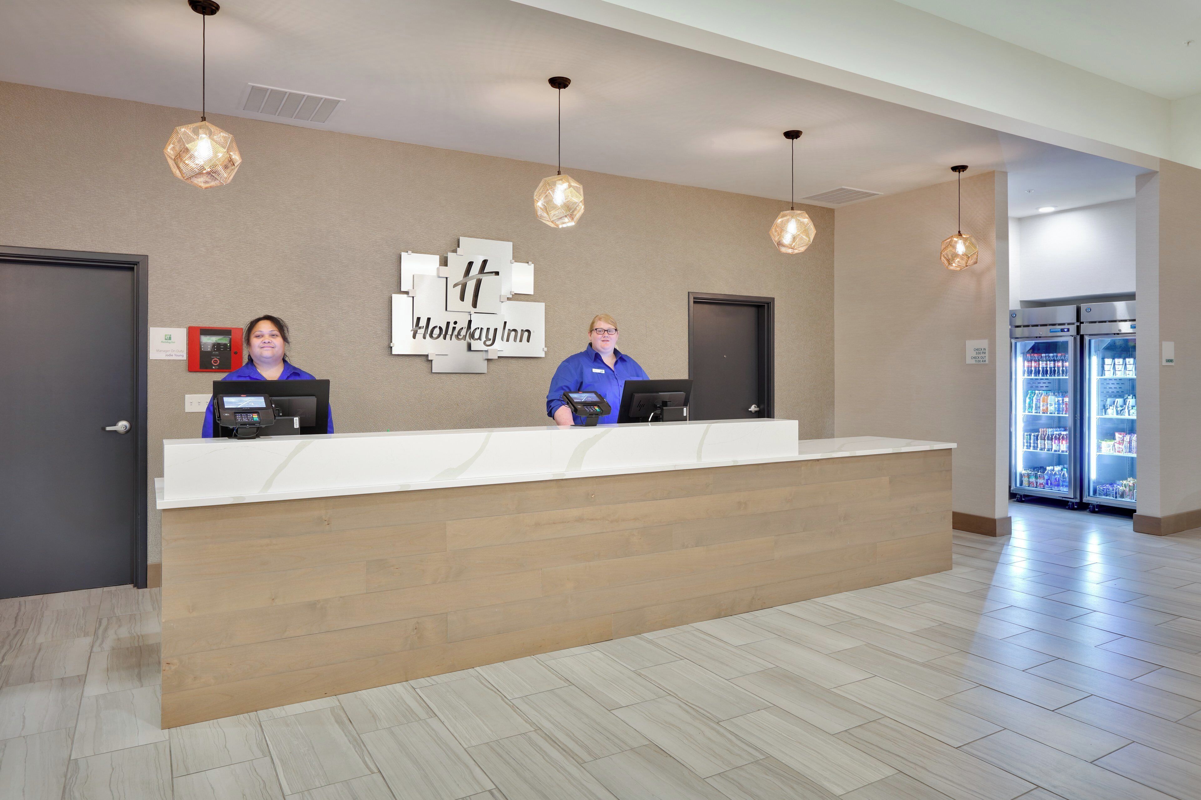 A reception desk with two staff members, a Holiday Inn sign, and a refrigerator stocked with beverages.