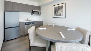A modern kitchen with grey cabinets, a stainless steel refrigerator, and a microwave. There is a dining table with four chairs and a framed picture on the wall.