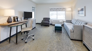 A modern hotel room with a flat-screen TV, a desk with a lamp, a grey armchair, a sofa, and a small ottoman. - Holiday Inn Twin Falls by IHG (Twin Falls)