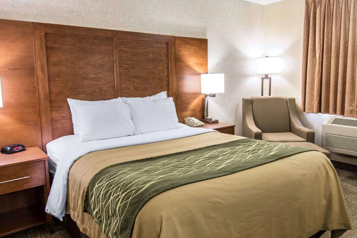 A hotel room with a large bed, a chair, a desk, a lamp, and an air conditioning unit.