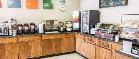Un área de desayuno autoservicio con máquinas de café, un reloj y contenedores de basura etiquetados.