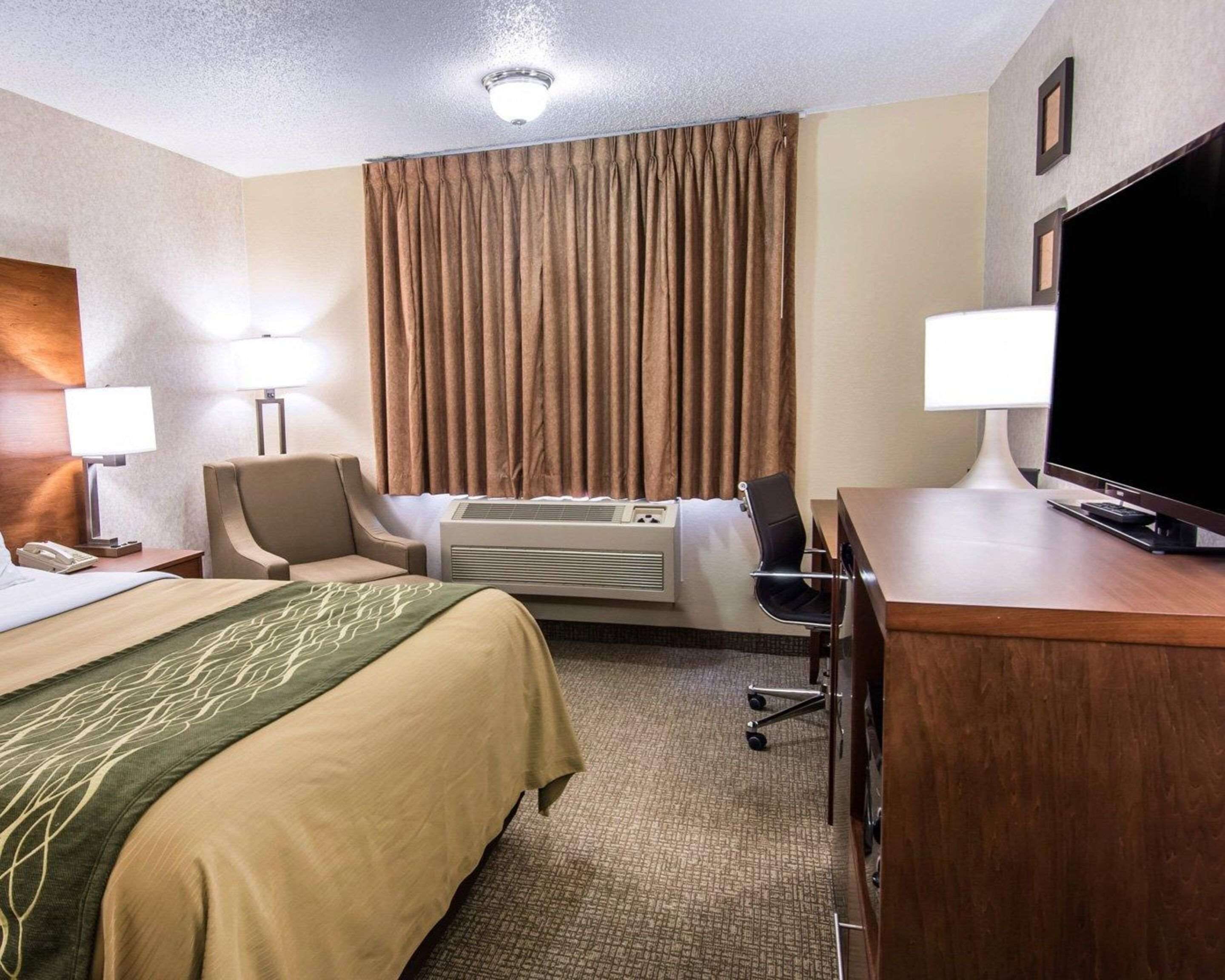 Quarto de hotel com cama, escrivaninha com televisão, cadeira, luminária de mesa, ar condicionado e telefone.