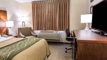 Quarto de hotel com cama, escrivaninha com televisão, cadeira, luminária de mesa, ar condicionado e telefone.