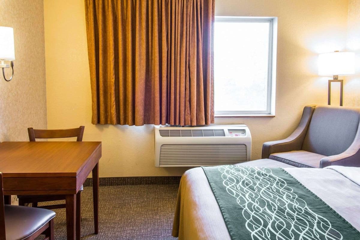 A hotel room with a bed, a desk, a chair, an air conditioning unit, a window with curtains, and a lamp.