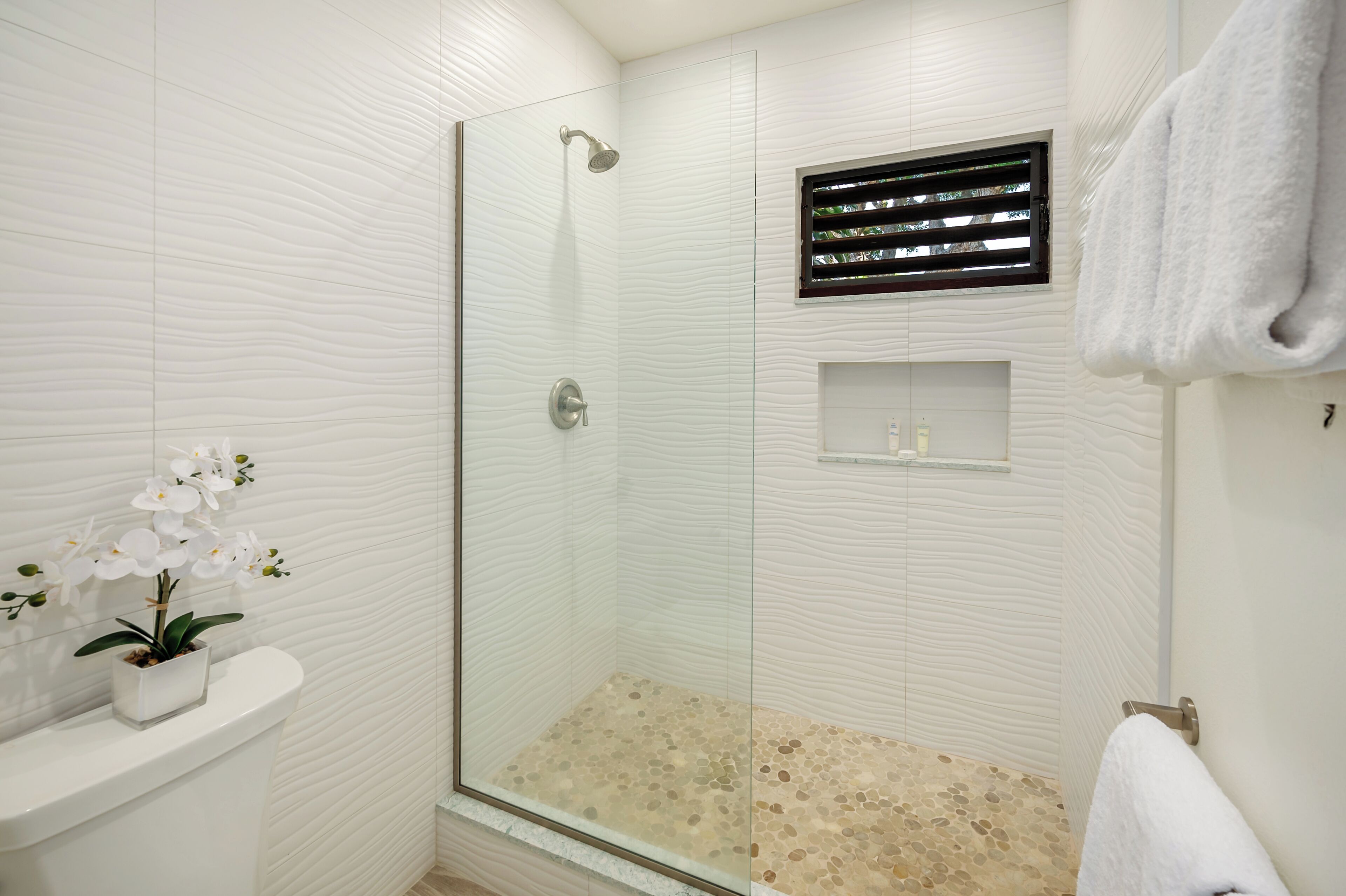 Une salle de bain moderne avec une douche en verre, un lavabo blanc orné d’une orchidée en pot et une serviette blanche accrochée au mur.