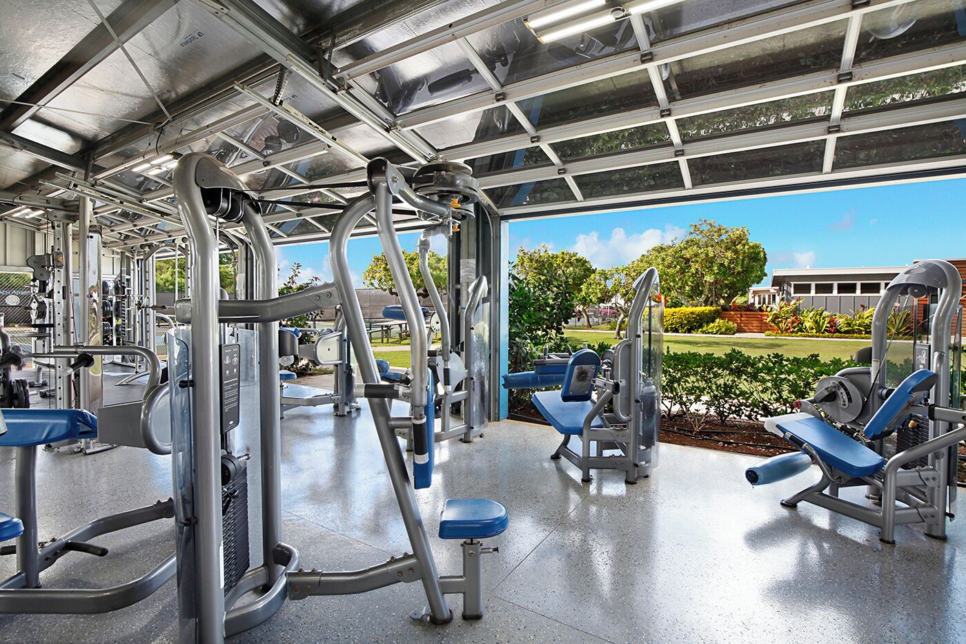A gym with various exercise machines, a covered area, and a view of a park and building.