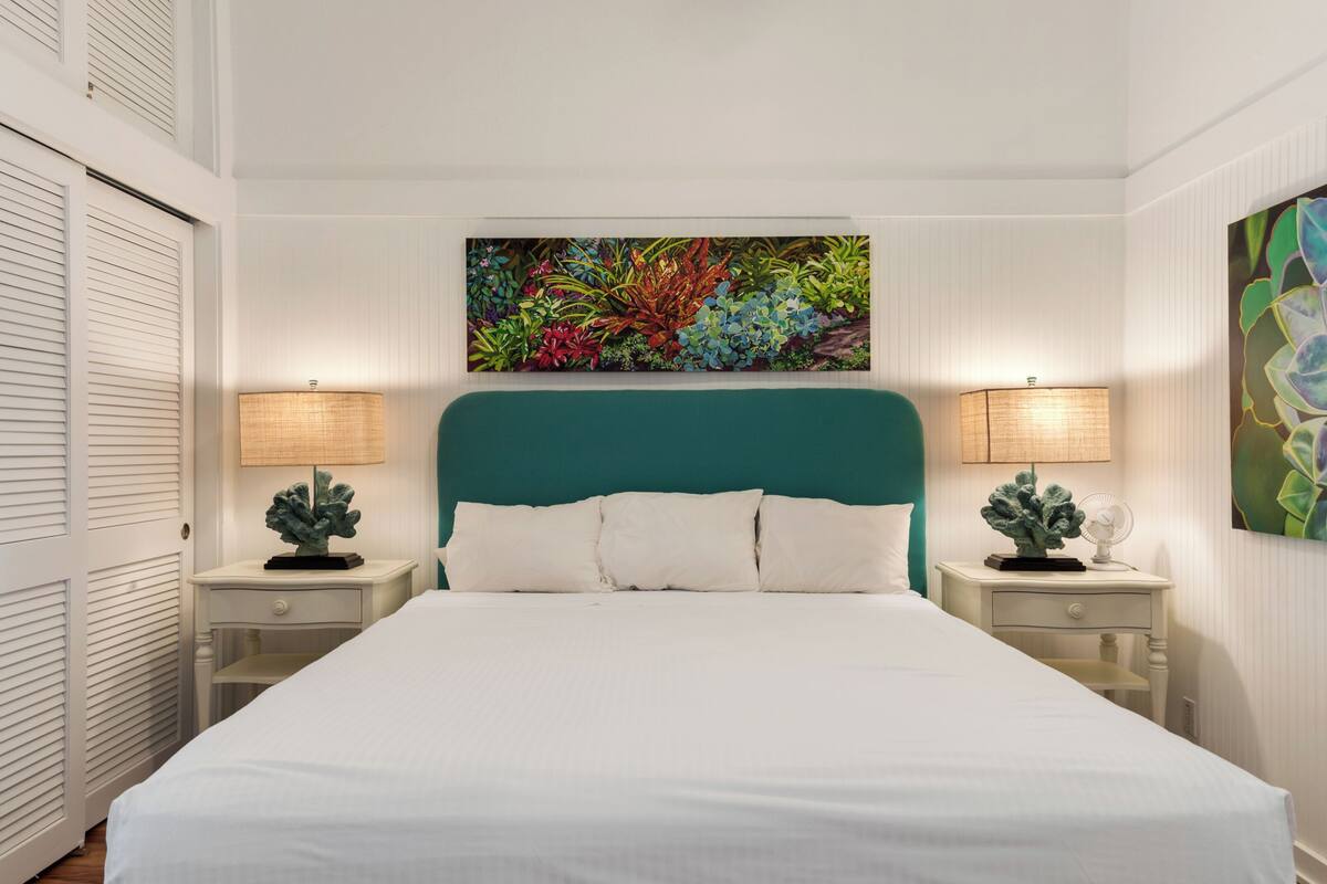 Ett hotellrum med en stor säng, två sängbord med lampor, en färgstark tavla ovanför sängen och en vit dörr till vänster.