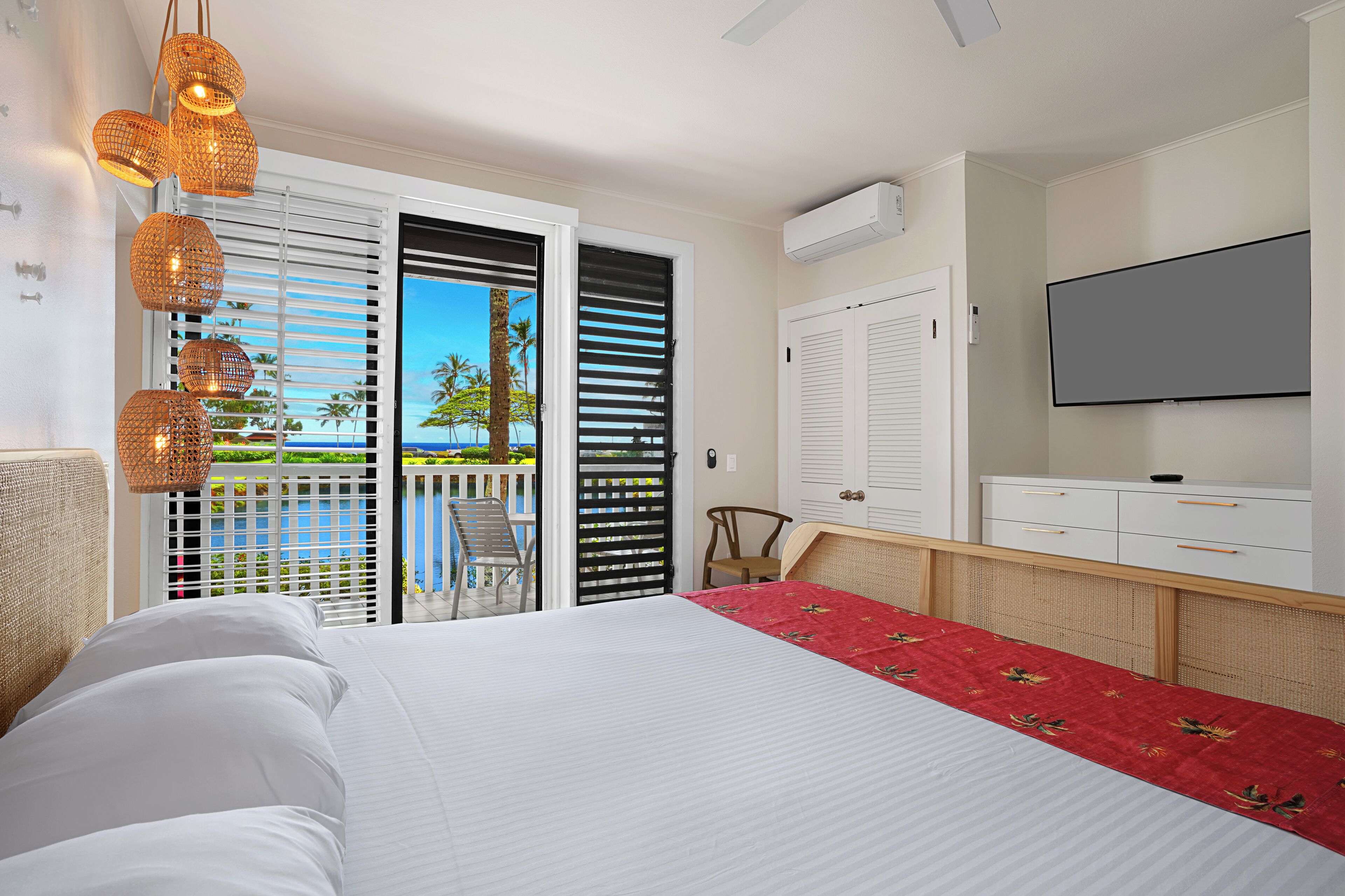 A hotel room with a bed, a TV, and a balcony with a view of palm trees and the ocean.