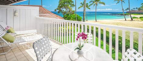 A covered outdoor seating area with white chairs and a table, overlooking a beach with palm trees and clear blue water.