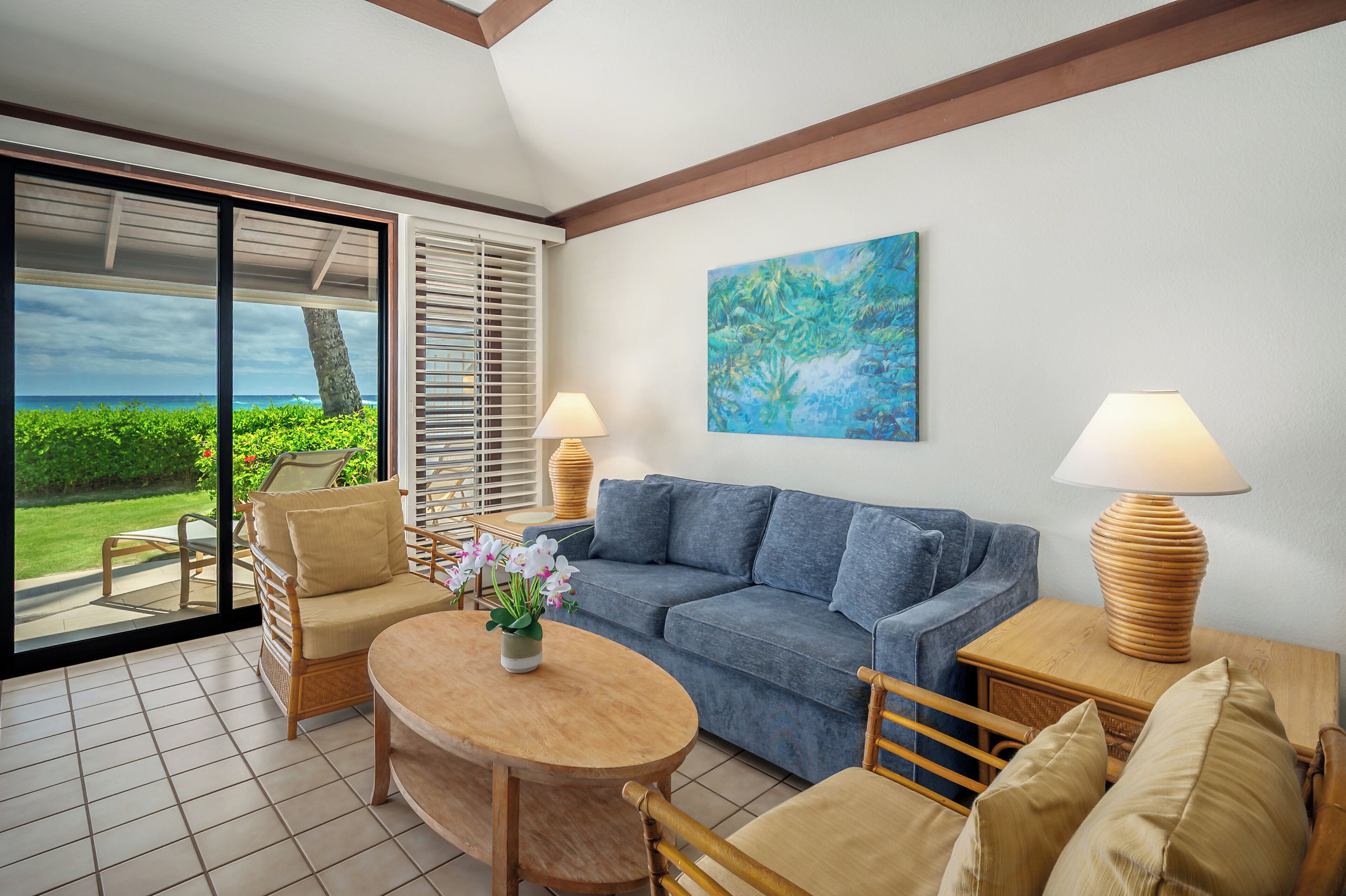 A modern living room with a blue sofa, wooden coffee table, and wicker chairs. There are two lamps on wooden side tables, and a painting on the wall.