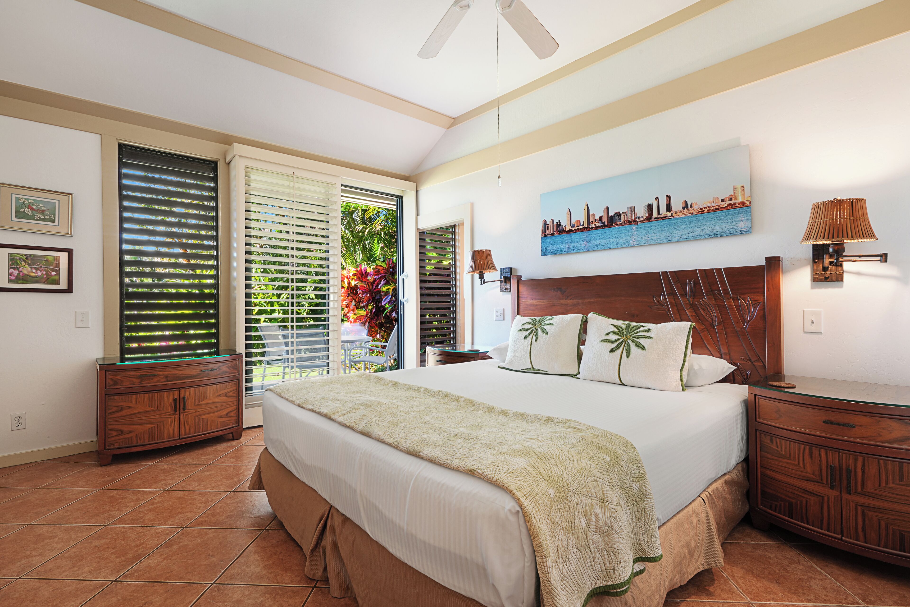 Una camera da letto con un letto grande, un quadro raffigurante un panorama urbano, una ventola a soffitto e vista su vegetazione dalla finestra.