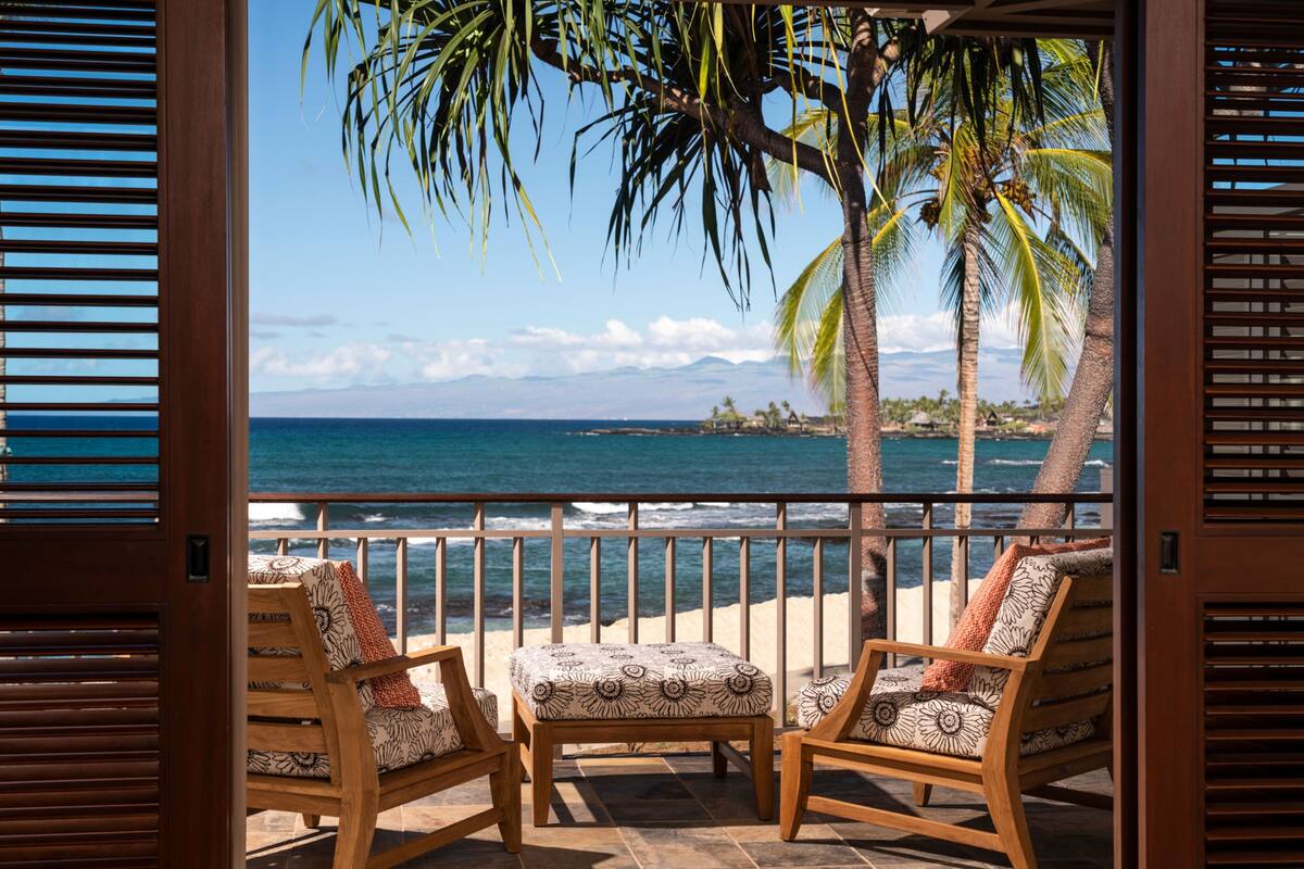 Un balcón de madera con dos sillas y una mesa pequeña, con vistas a una playa con palmeras y el océano.