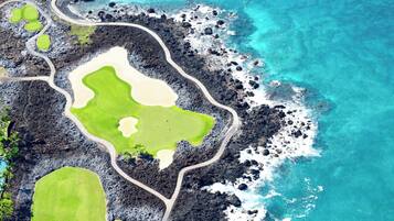 An aerial view of a coastal golf course with a winding road, green fairways, and a sandy beach area.