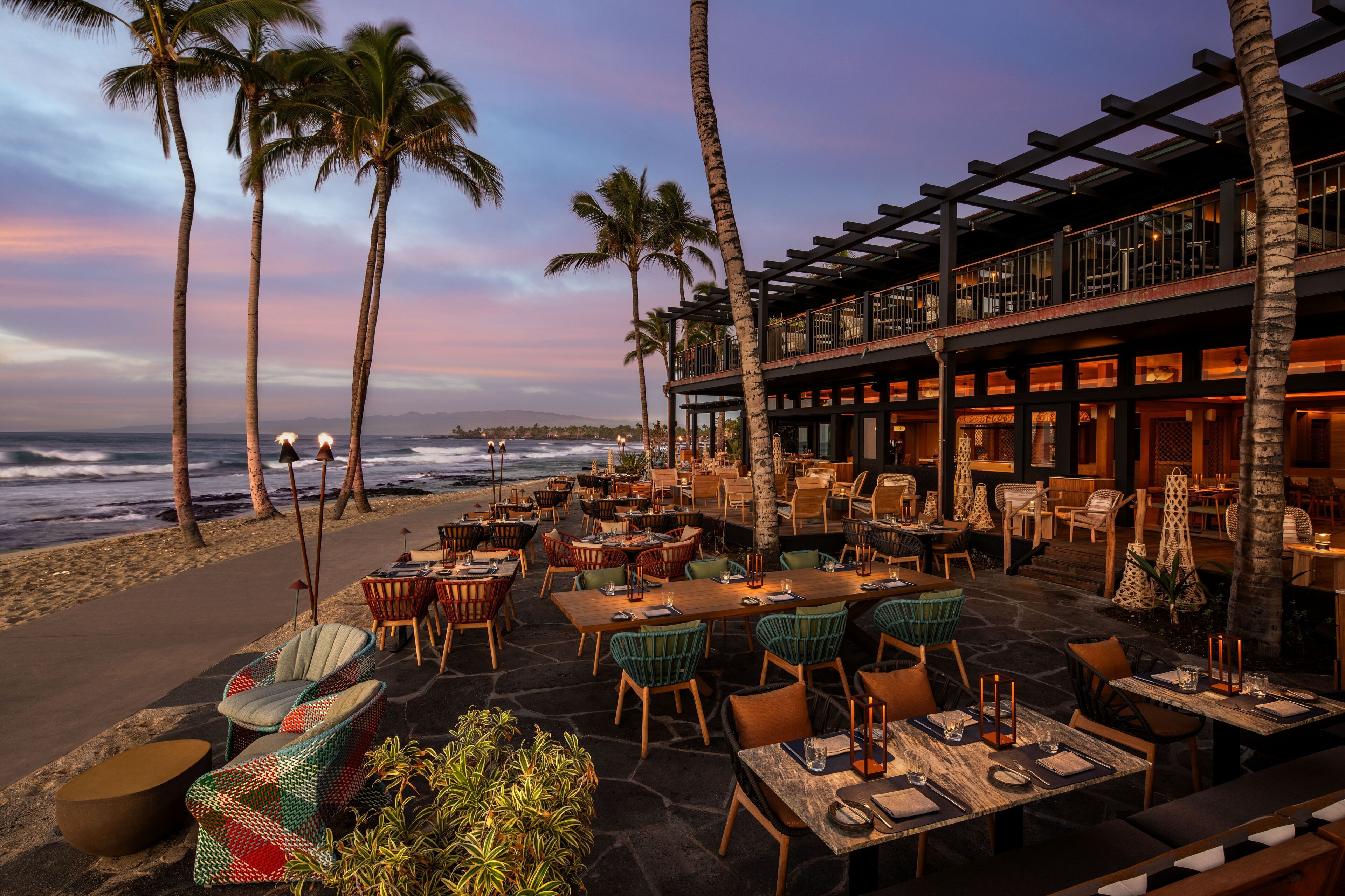 An outdoor restaurant with tables and chairs, palm trees, and a view of the ocean.