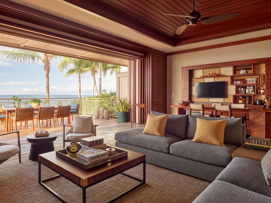 A spacious living area with a large sofa, coffee table, and a TV mounted on the wall. There is a balcony with outdoor seating and a view of the ocean.
