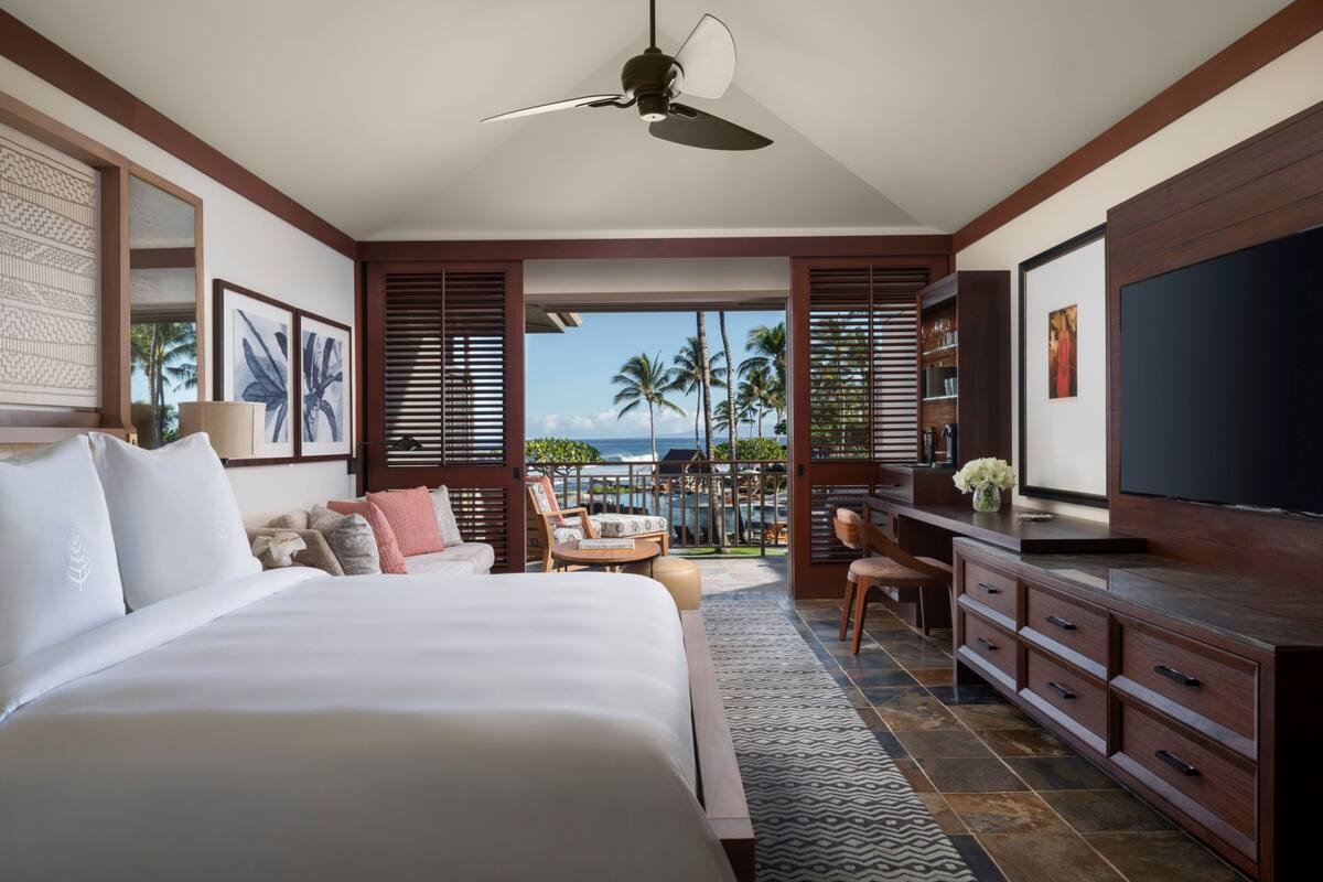 Habitación de hotel con una cama grande, televisión, escritorio y vistas a palmeras y al océano.