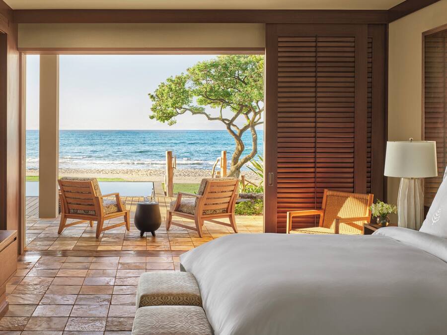 A hotel room with a large bed, a TV, a wooden floor, a view of the beach, and a dining area with chairs and a table.