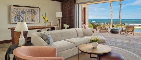 A modern living room with a large sofa, a coffee table, and a chair. There is a view of the ocean through large windows.