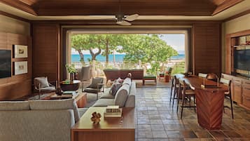 A spacious living area with a view of the ocean, featuring a sofa, chairs, a dining table, and a ceiling fan.