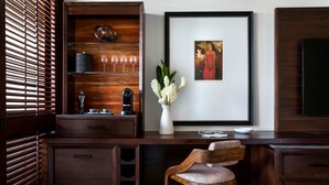 A wooden desk with a chair, a vase with flowers, a framed picture, and a shelf with wine glasses and a clock.
