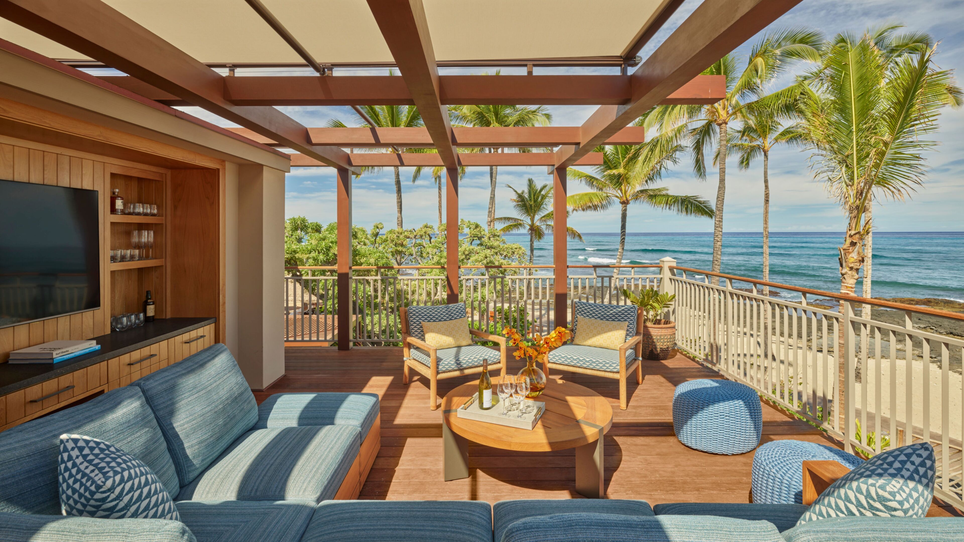A covered outdoor seating area with a wooden deck, a sofa, two armchairs, and a coffee table. There is a TV on a wooden cabinet to the left. The area overlooks a beach with palm trees and the ocean.