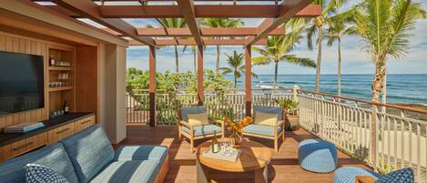 A covered outdoor seating area with a wooden deck, a sofa, two armchairs, and a coffee table. There is a TV on a wooden cabinet to the left. The area overlooks a beach with palm trees and the ocean.