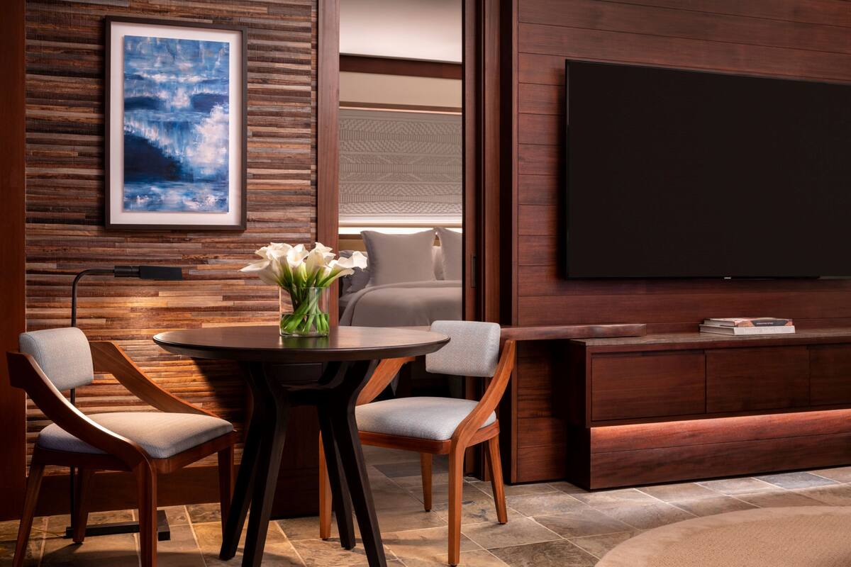 Una sala moderna con una mesa redonda de madera, dos sillones tapizados, un televisor de pantalla plana grande, un cuadro abstracto enmarcado y un mueble empotrado para entretenimiento.