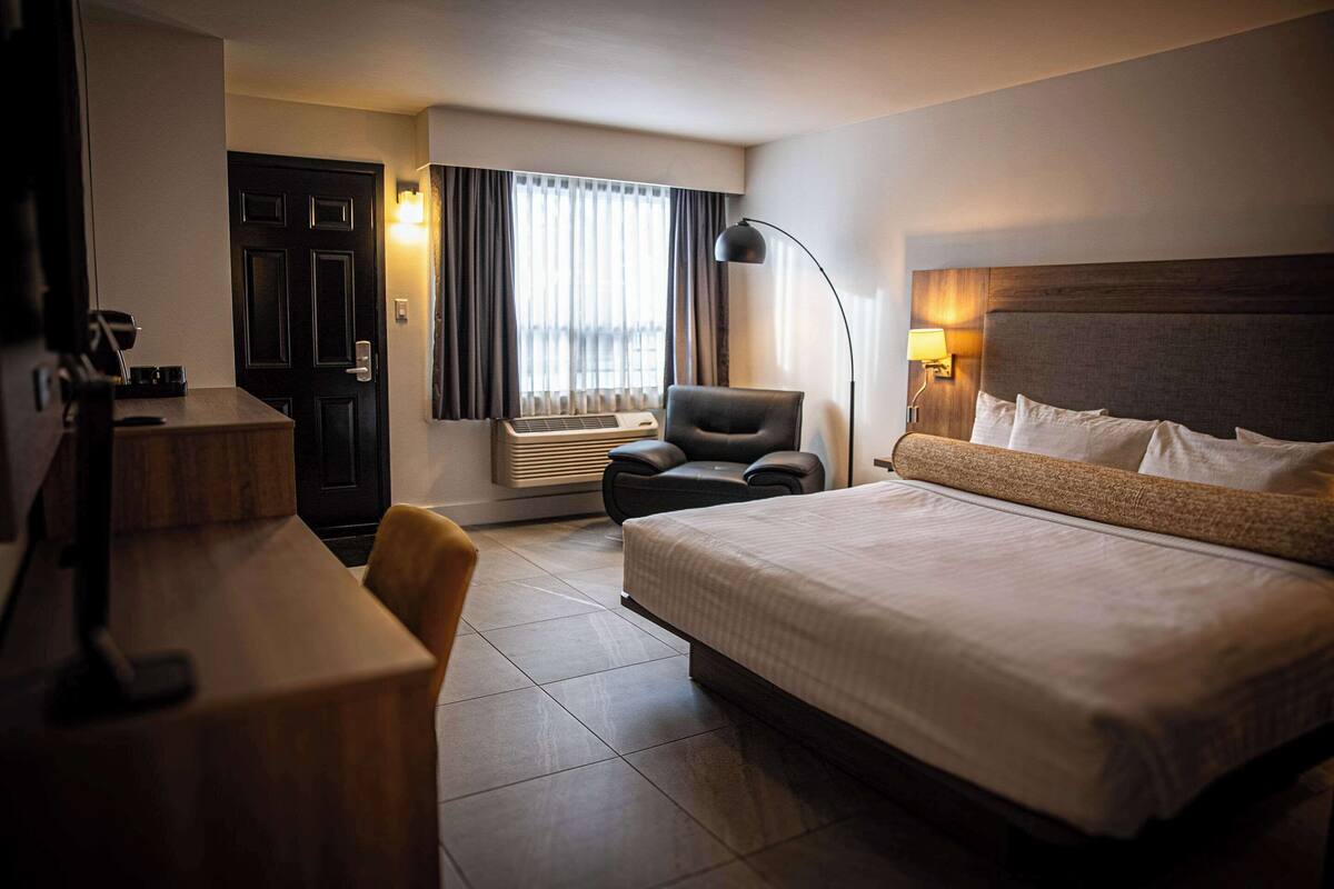 A hotel room with a large bed, a desk with a chair, a desk lamp, a chair, a window with curtains, and an air conditioning unit.