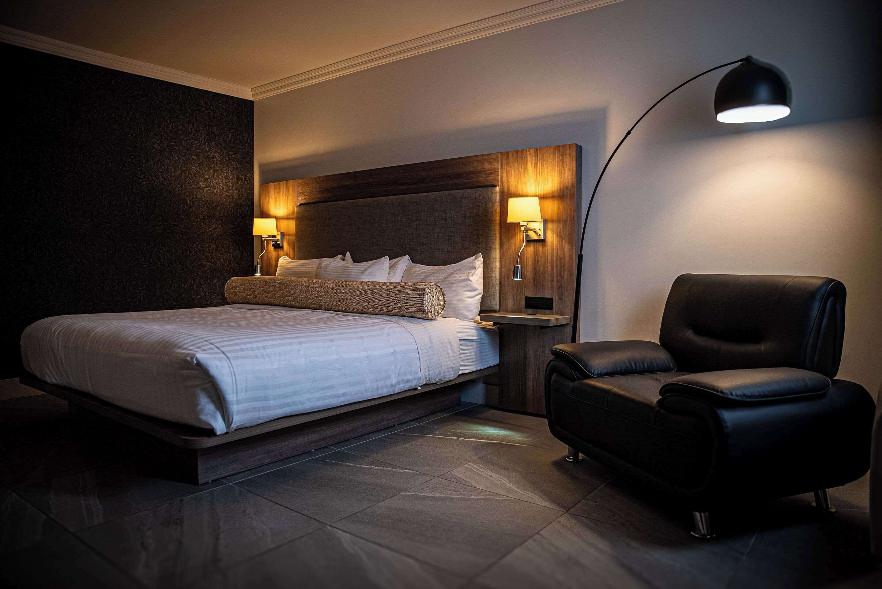 A modern hotel room with a large bed, two bedside lamps, a black leather chair, and a dark textured wall.