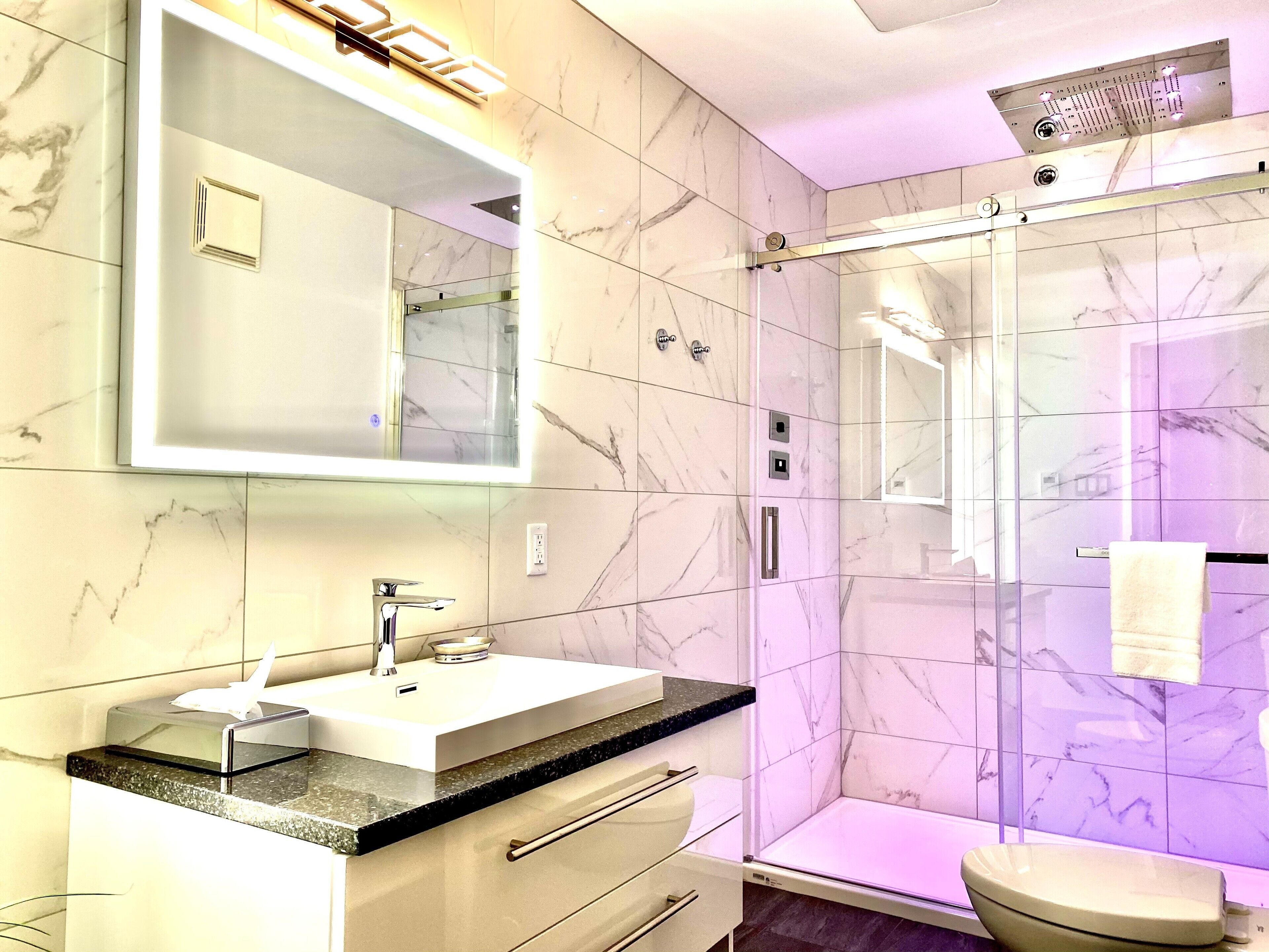 Un baño moderno con pared de azulejos de mármol, ducha con mampara de vidrio, lavamanos blanco con encimera negra y un espejo grande encima.