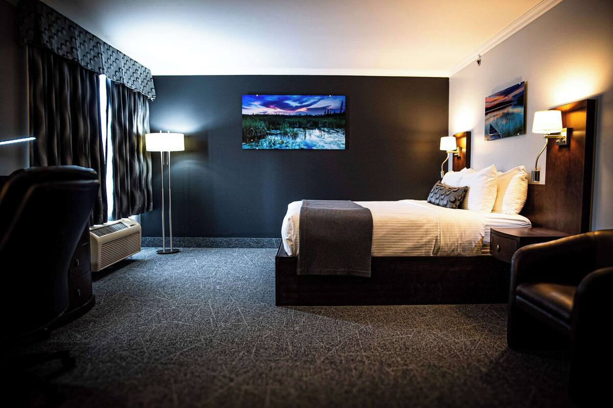 A hotel room with a bed, two bedside lamps, a wall-mounted painting, a floor lamp, and a chair.