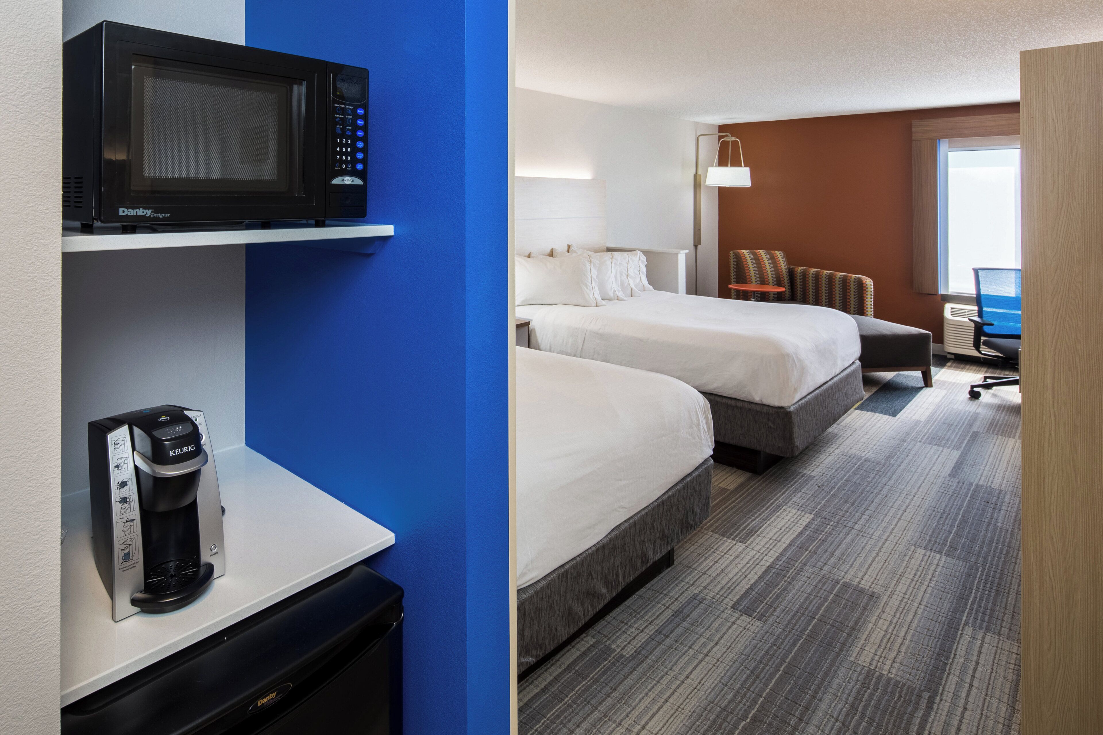A hotel room with a microwave, coffee maker, two beds, a desk, a chair, and a view of the outdoors.