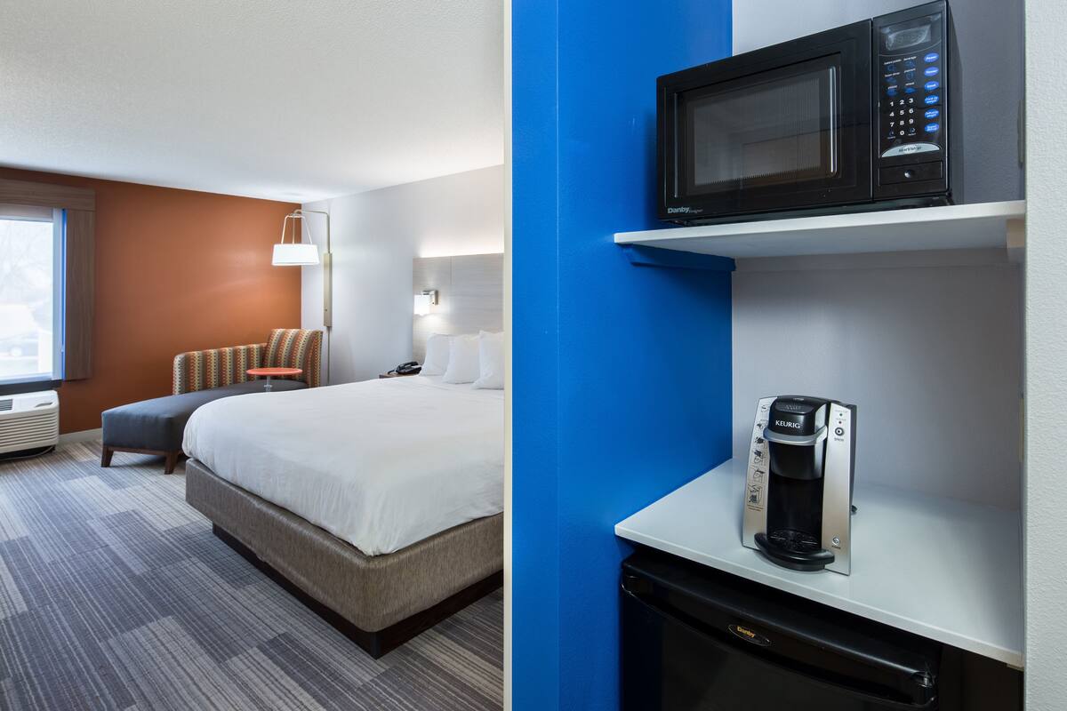A hotel room with a bed, a desk with a microwave and coffee maker, a lamp, and a window with curtains.