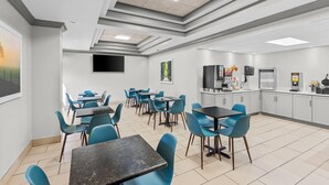 A modern dining area with blue chairs and black tables, a flat-screen TV mounted on the wall, and a kitchen area with appliances and a refrigerator. - Days Inn & Suites by Wyndham Cedar Rapids (Cedar Rapids)