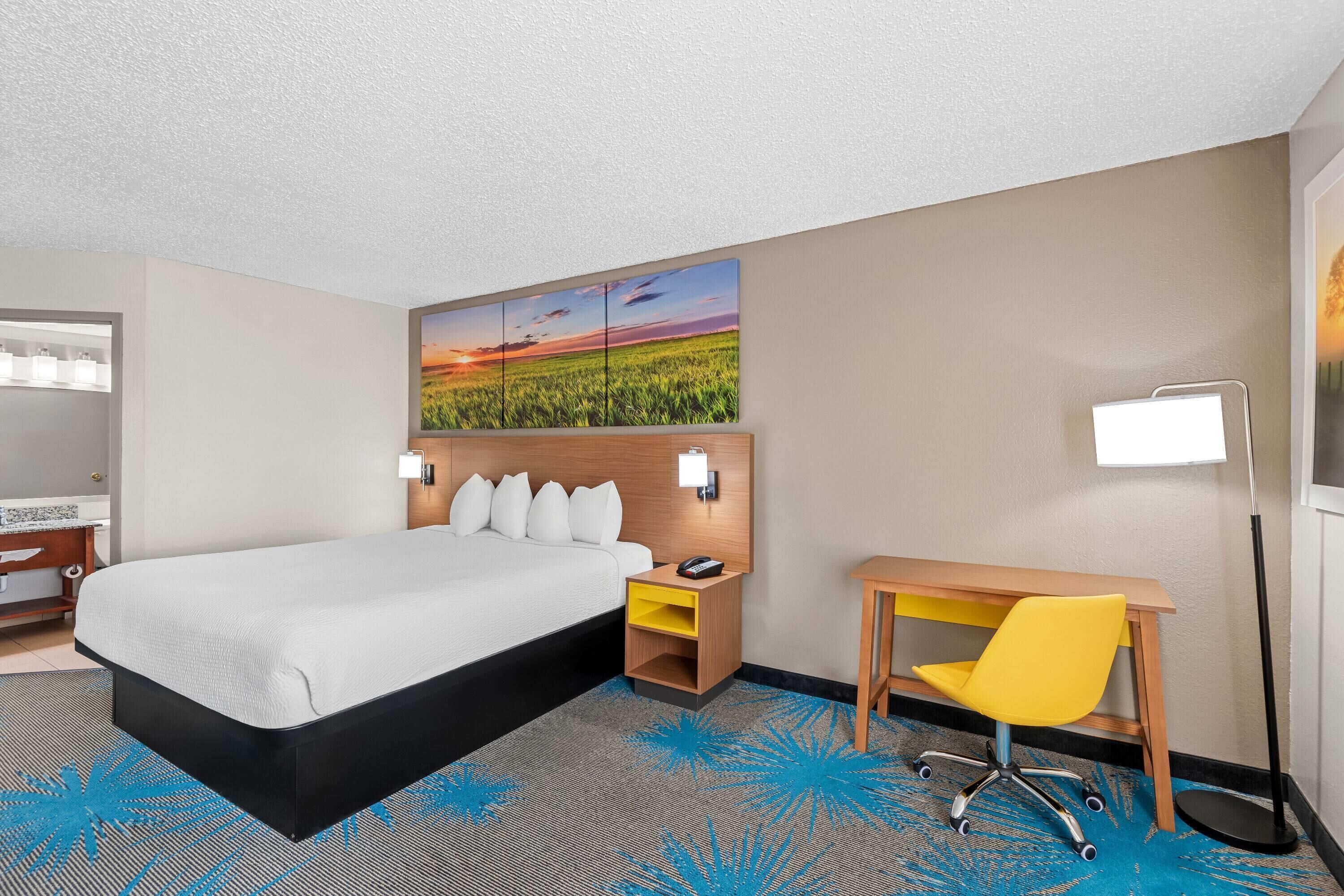 A hotel room with a bed, a desk, a chair, a nightstand, a lamp, and a wall art piece.