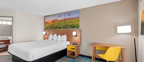 Una habitación de hotel con cama, escritorio, silla, mesita de noche, lámpara y una obra de arte en la pared.