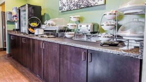 Montaje de bufé con distintas estaciones de comida, que incluyen una máquina de café, una cesta de frutas y varios platos para servir.