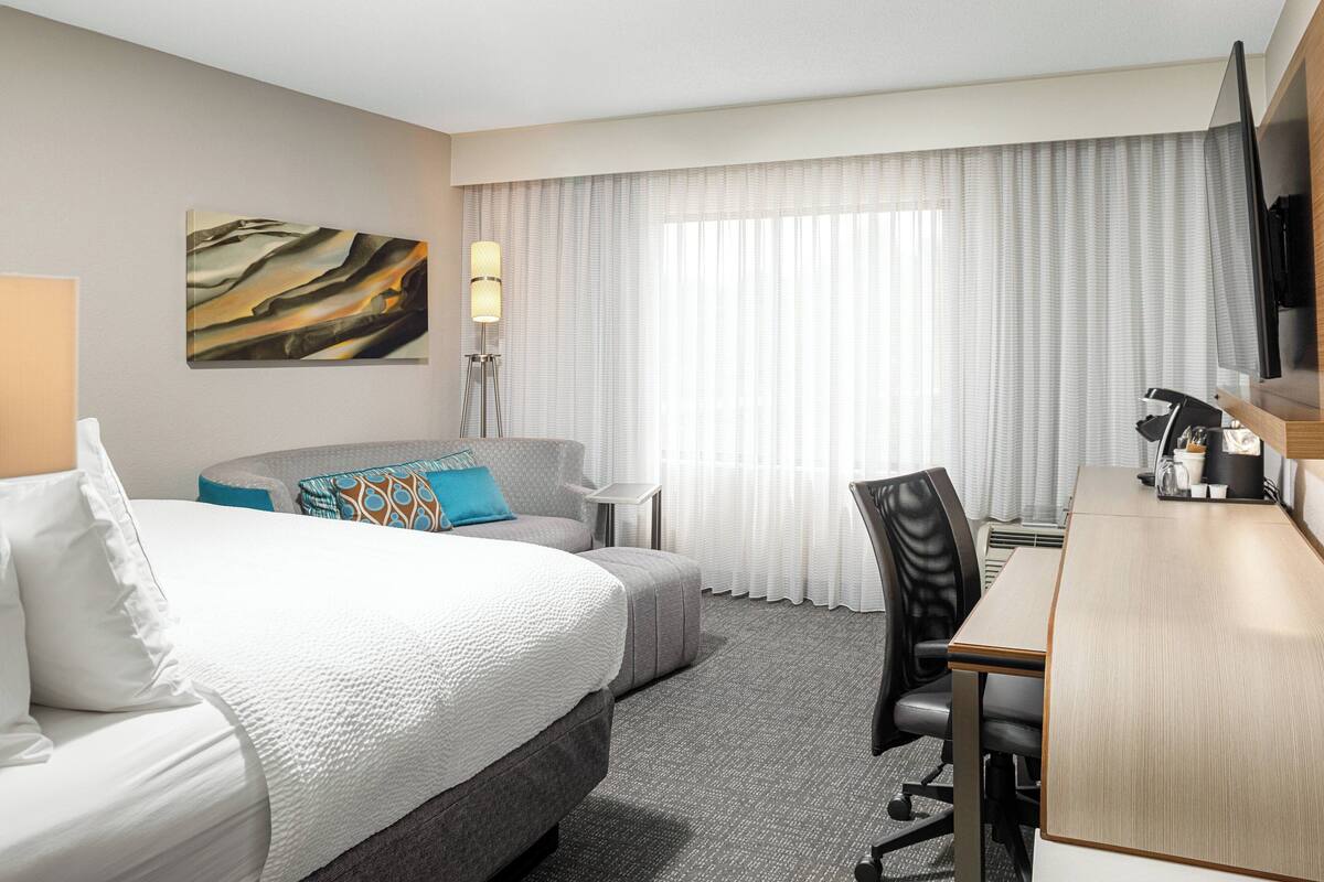 A hotel room with a bed, a desk, a chair, a TV, and a painting on the wall.