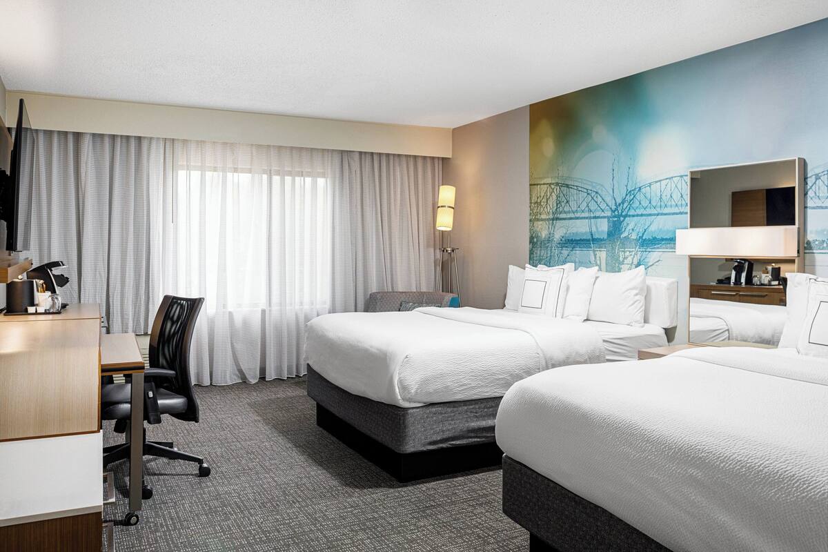 A hotel room with two beds, a desk, a chair, a lamp, and a painting of a bridge on the wall.