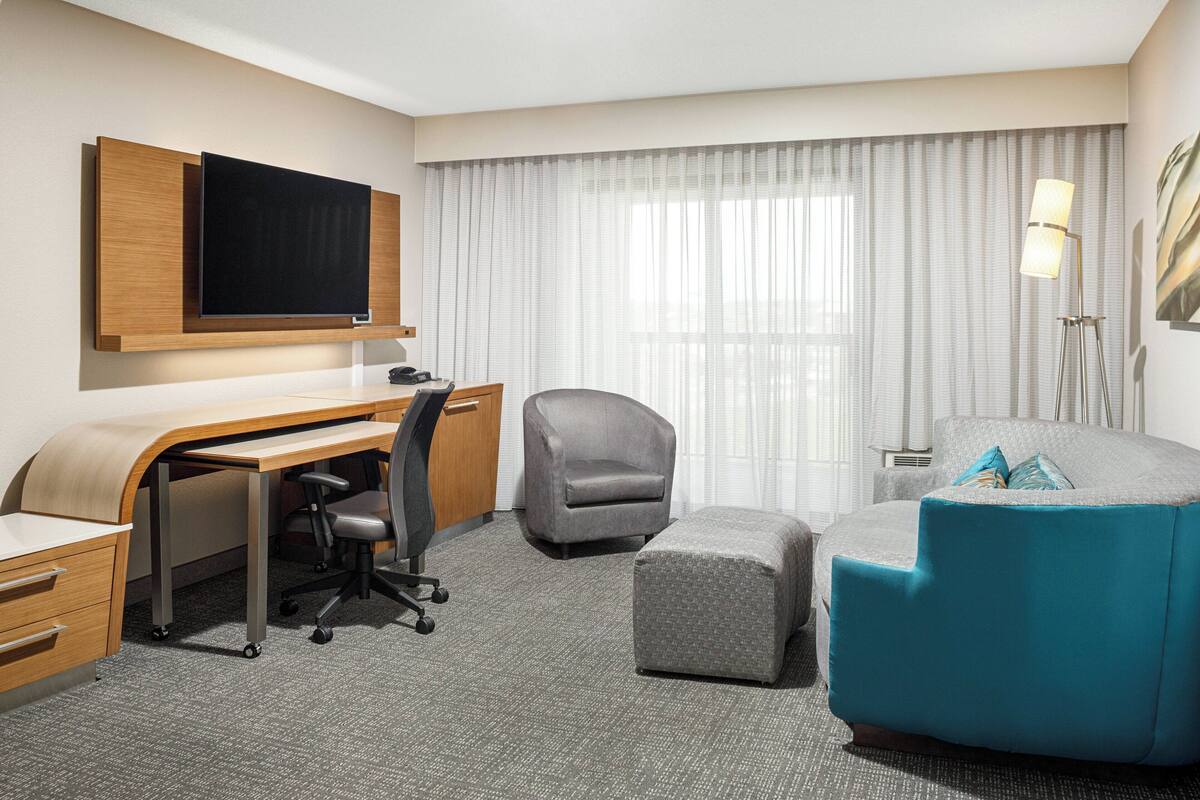 A hotel room with a desk, a grey armchair, a blue sofa, and a television mounted on the wall.