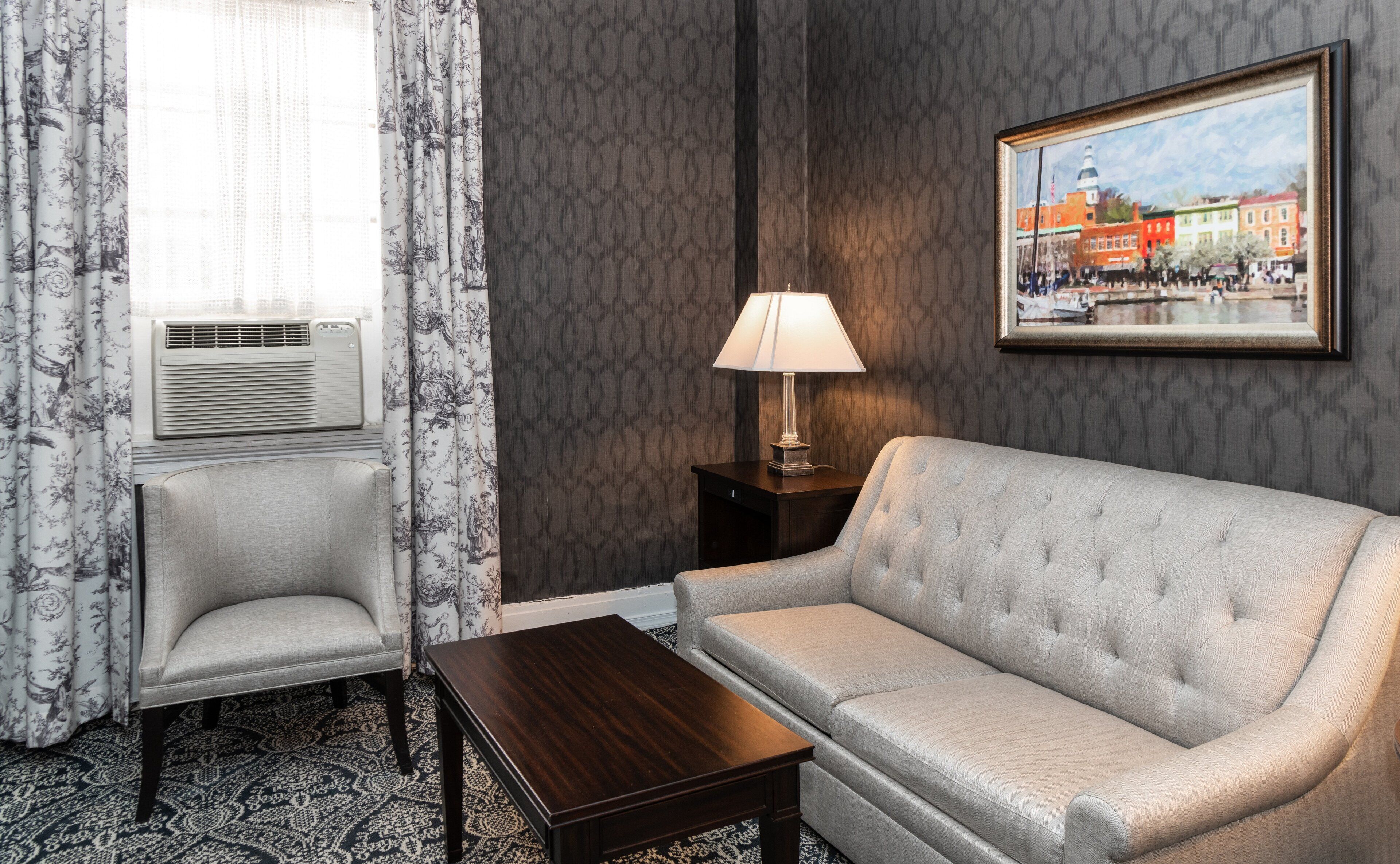 A hotel room with a grey sofa, a small armchair, a wooden coffee table, a lamp on a nightstand, and a painting of a cityscape on the wall.